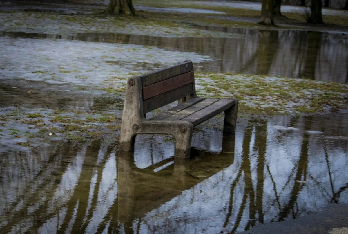 Wooden bench mirrored in floodwater across a muddy park lawn